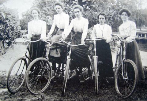 black and white photograph of five individuals in a row wearing white blouses and dark skirts holding or sitting on bicycles outdoors