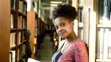 Smith looking at camera, holding open book, leaning against bookshelf in library stacks