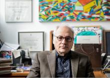 Charles Bernstein at his desk with a colorful painting by his wife hanging on wall behind him