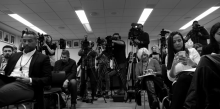 Journalists sitting on chairs on their phones in front while others man tripod-supported video cameras, in black & white