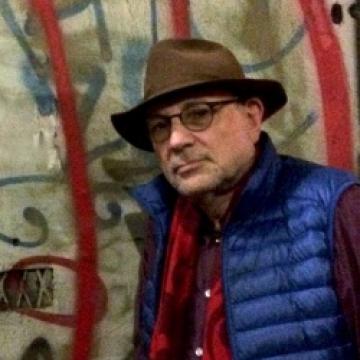 Charles Bernstein, wearing a red shirt, blue vest, and brown hat, stands in front of colorful art