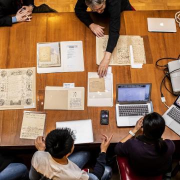 Session of Manuscript Collective session, where students and Peter Stallybrass examine documents on a wooden table in the Lea Library
