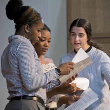 Three students stand together, speaking while consulting printed papers