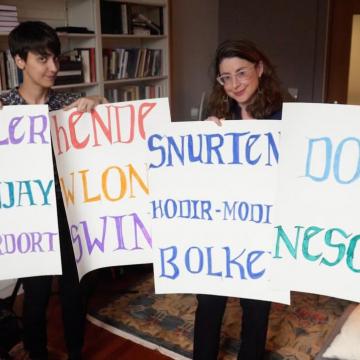 From video: Emily Steiner and Aylin Malcolm in academic office in front of bookcases hold up four white posters with 11 hand-lettered words written in  bright-colored marker
