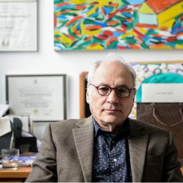 Charles Bernstein at his desk with a colorful painting by his wife hanging on wall behind him