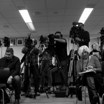 Journalists sitting on chairs on their phones in front while others man tripod-supported video cameras, in black & white