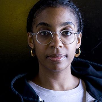 Headshot of Simone White wearing a white shirt, black hoodie, glasses, and gold earrings.